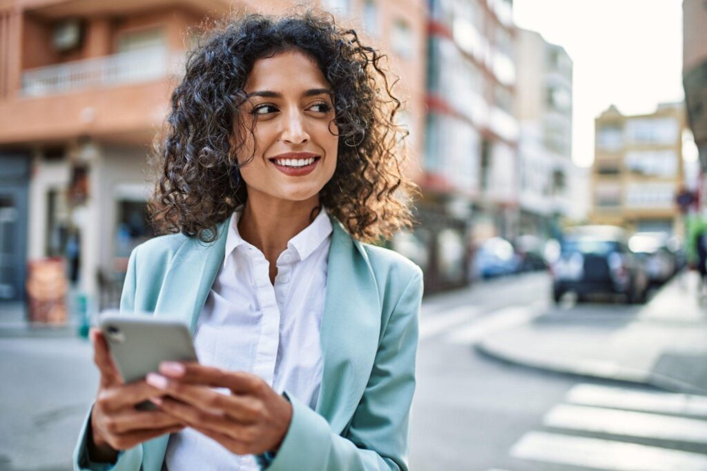 Mujer hispana joven del negocio que usa mirada profesional que sonríe confidente en la ciudad usando el smartphone