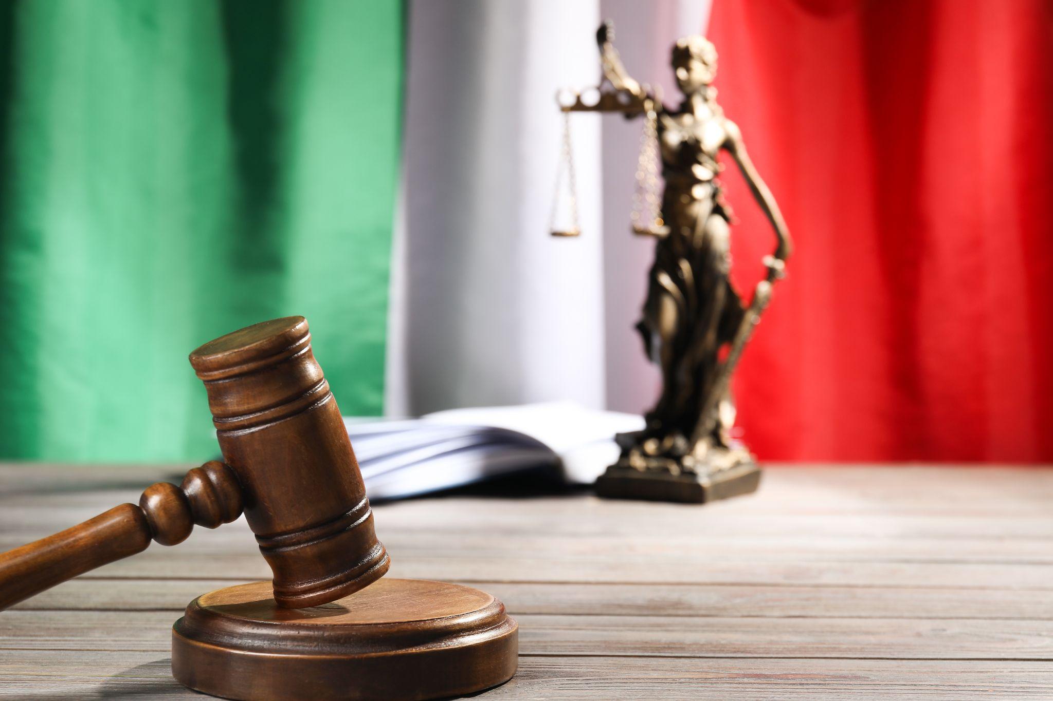 A legal gavel and a lady justice statue on a table with an Italian flag in the background