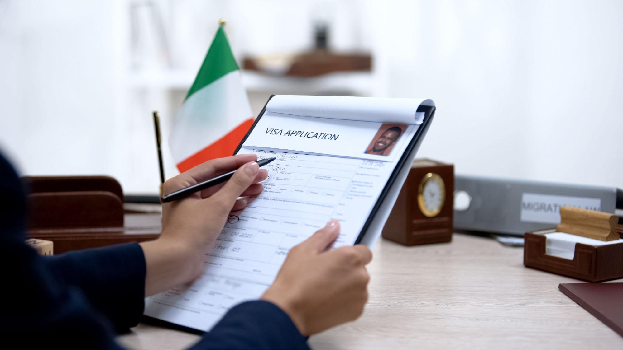 Hands holding a visa application with an Italian flag on his desk